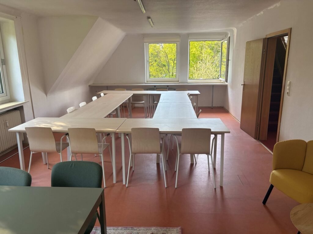 Seminarraum unter dem Dach mit Tischen in U-Form, grünen Stühlen und gelbem Sessel